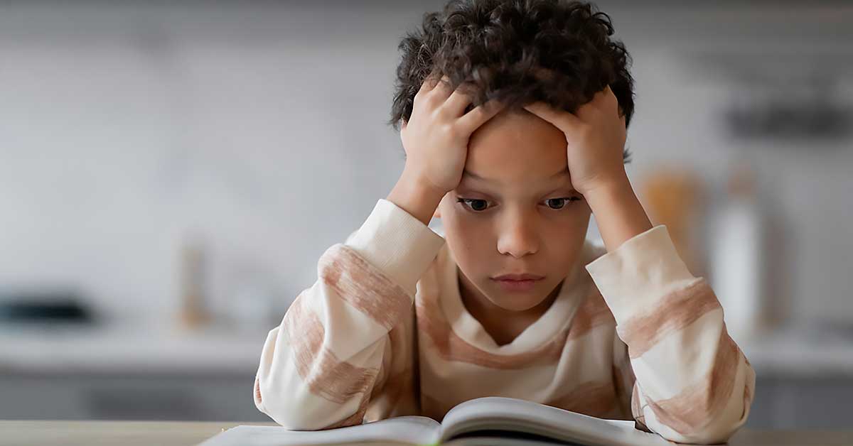 Stressed Black second grade boy reading a book. His expression is stressed and he is holding his hands to his head.